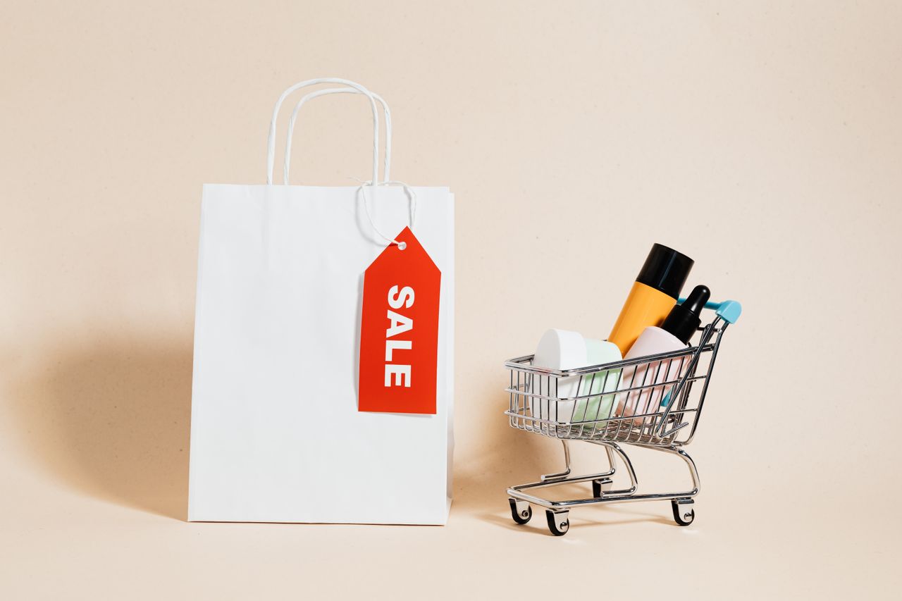 White shopping bag with red sale tag and mini shopping cart on beige background.